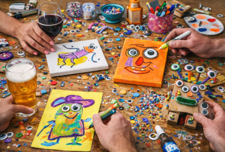 Adults enjoying a quirky craft night, painting funny faces on small canvases at a messy table with colorful craft supplies and drinks for a playful, creative evening at home.